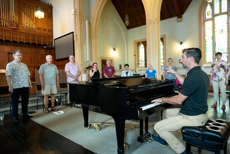 Members of the choir practice practicing while Roland plays piano.